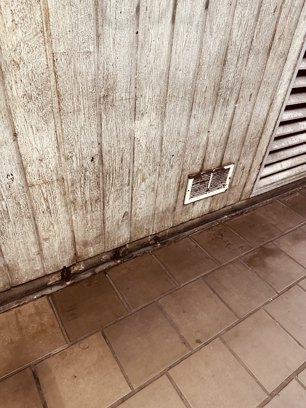 A stained board-formed concrete wall beside an HVAC vent grille, with grime along the baseboard and tile floor.
