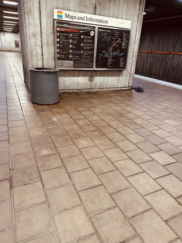 A Maps and Information board mounted on a board-formed concrete wall above a metal trash can and stained tile floor.