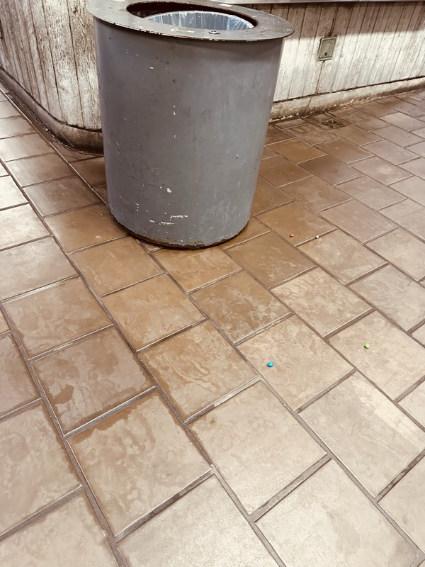 Close-up of a scarred, scratched metal trash can sitting on a heavily soiled tile floor.