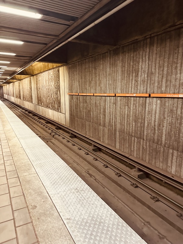 Arts Center platform viewed from the track edge, showing decorative wall panels and an orange Arts Center strip running along weathered concrete.
