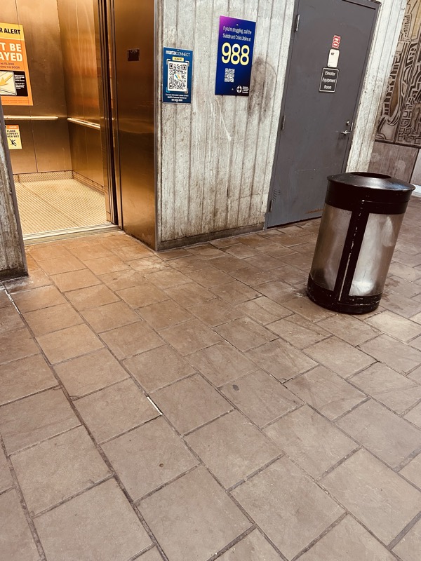 A 988 mental-health crisis-line sign mounted beside an elevator and a metal trash can, set against a weathered board-formed concrete wall and a stained tile floor.