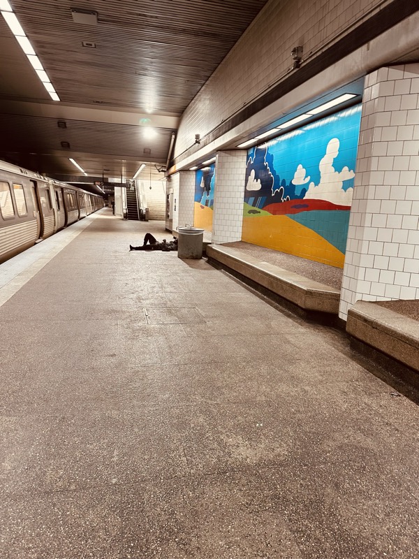 A person rests on the ground next to a colorful public-art mural and a concrete bench on a station platform.