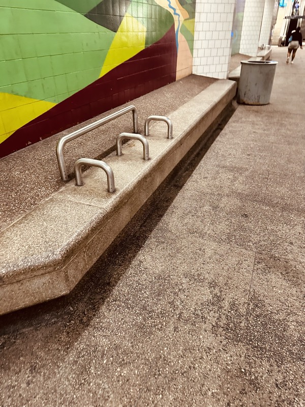 Anti-loitering metal bars installed along a concrete bench beside a station mural, with a soiled granite floor in front.