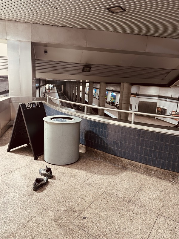 A pair of sneakers left on the floor beside a concrete trash receptacle and a folded A-frame sign on a station mezzanine.