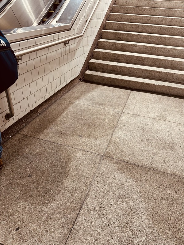 A wide concrete landing at the base of an escalator and stairwell, with white tile walls grayed by years of grime.