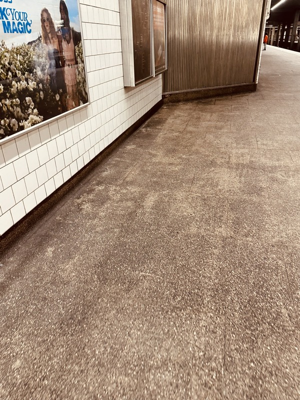 A long stretch of stained concrete station floor running alongside a white-tile wall with a bright wellness advertisement.