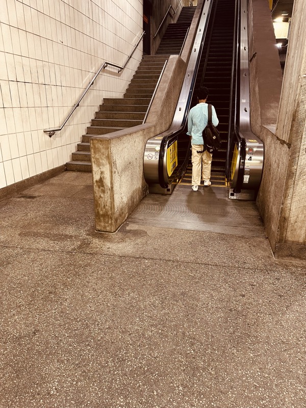 A commuter rides an upward escalator. The concrete floor at the base of the escalator is darkened almost to black with accumulated grime.