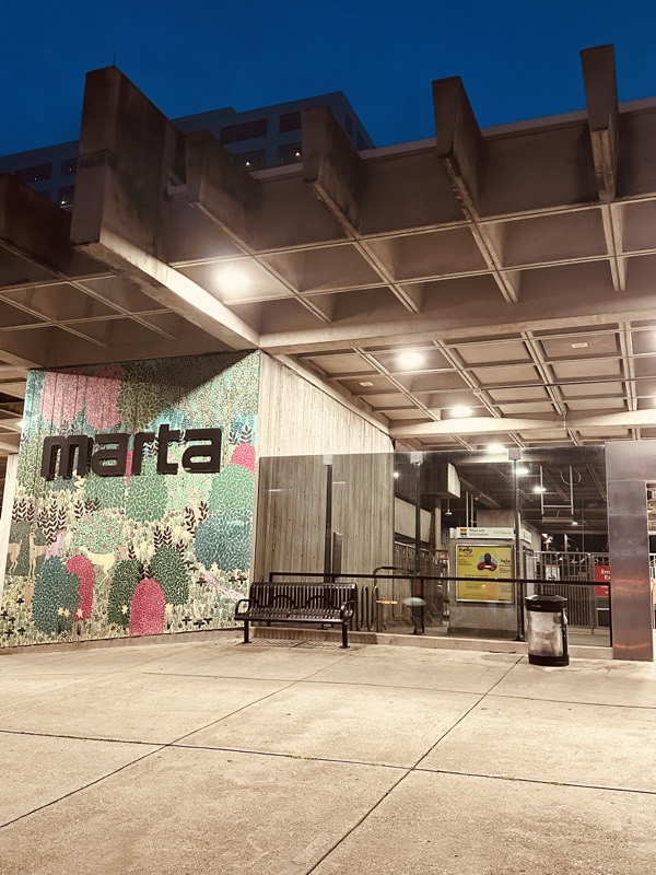 The exterior of a MARTA station at dusk, with its signature green-and-pink mural wall, station signage, and benches under a concrete canopy.
