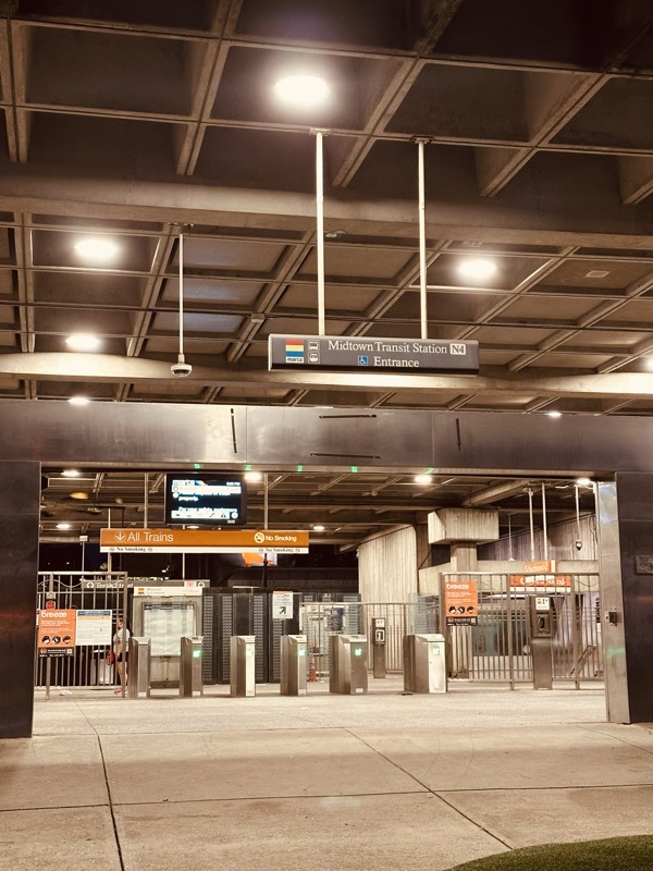 Midtown Transit Station entrance at night — the fare gate hall lit beneath an overhead concrete canopy.