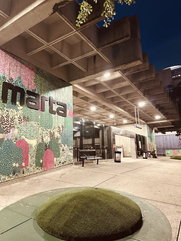 A MARTA station outdoor concourse at night with a large mural wall, benches, and a round AstroTurf installation underfoot.