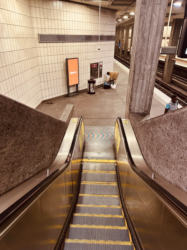 Looking down a station's downward escalator; a person crouches at the bottom near a trash bin and signage.