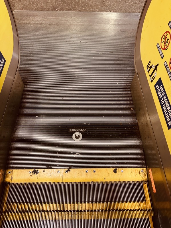 Looking straight down an escalator tread, showing scratches, paint marks and accumulated discoloration on the metal.