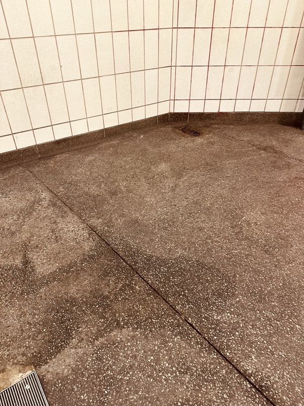 A station corner where a stained granite floor meets a white tile wall, the grout darkened to a rust line.