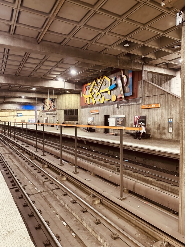 A wider view of Midtown station platform, looking across the tracks at a colorful abstract mural under a coffered concrete ceiling.